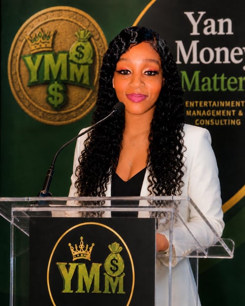 Woman in white blazer speaking at YMM podium with entertainment management branding on green backdrop